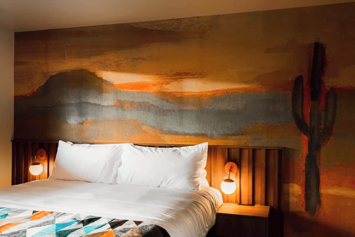 Una habitación de hotel con una cama, una mesita de noche con lámpara y un cuadro grande en la pared.