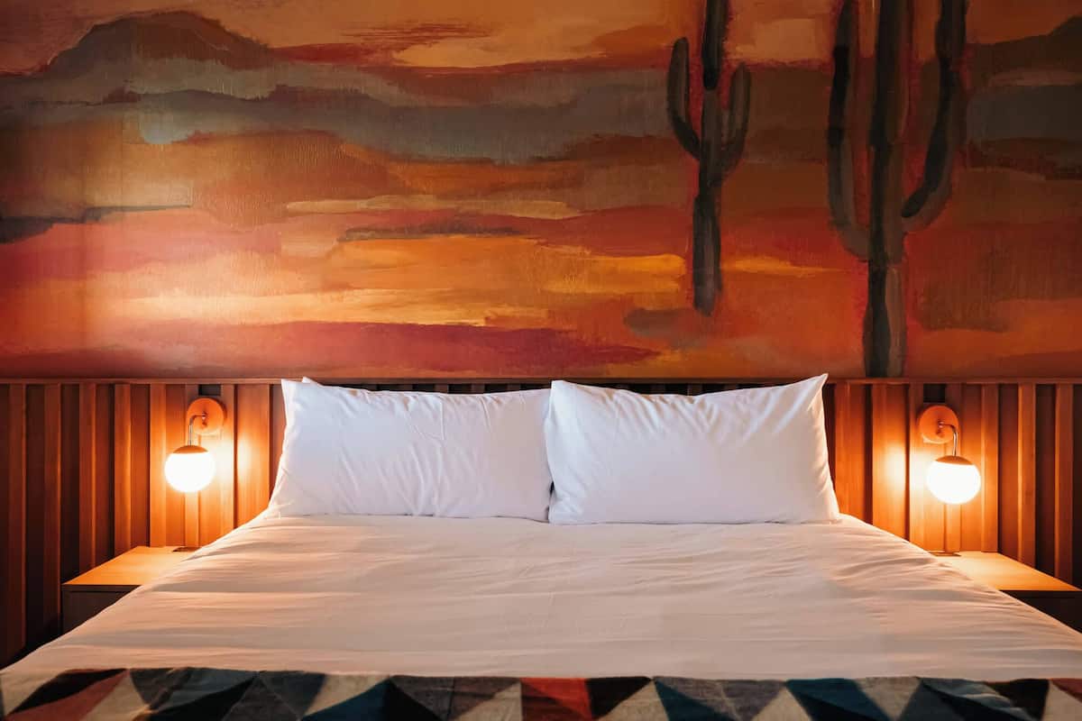 Una cama con sábanas blancas, dos lámparas de pared y un mural con un paisaje desértico que incluye un cactus.