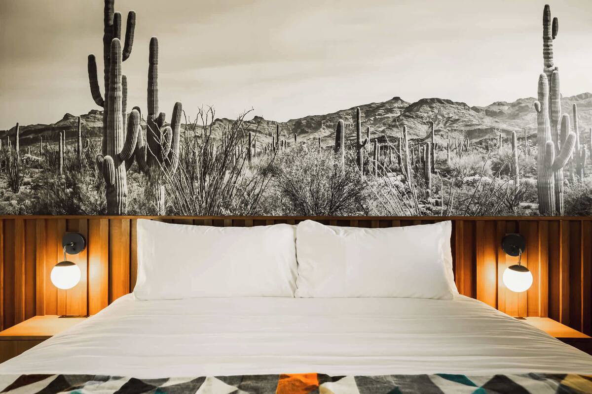 Una cama con sábanas blancas, dos lámparas de noche y un mural con motivos del desierto en la pared.