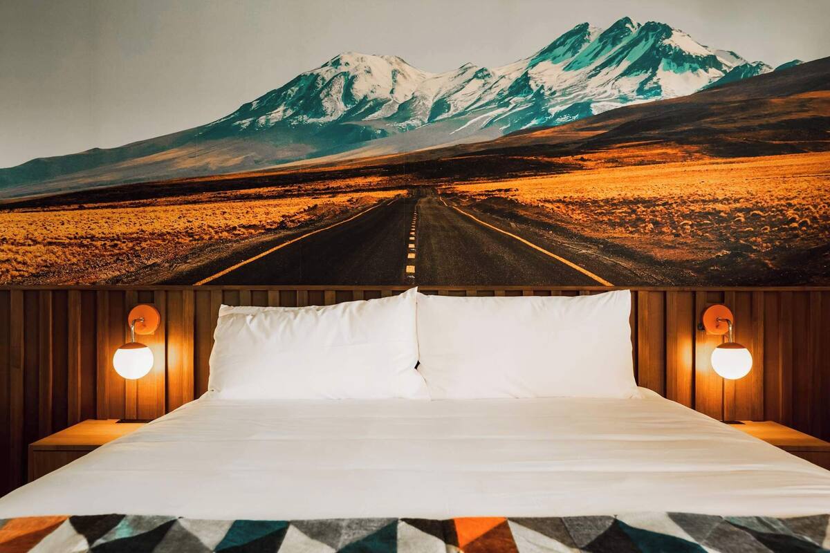 Una cama con un mural de vistas a la montaña, cabecera de madera y dos lámparas fijadas a la pared.