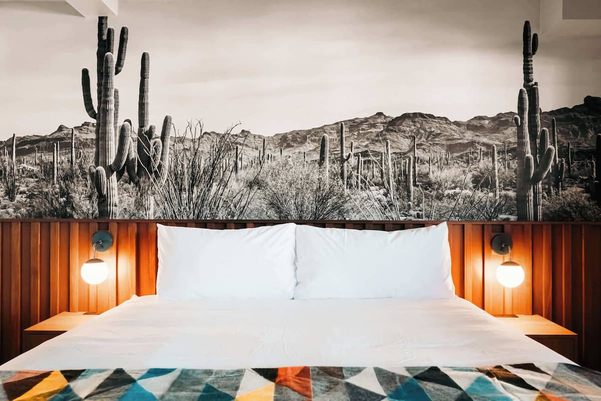 Habitación de hotel con cama, almohadas blancas y un mural grande con motivos del desierto.