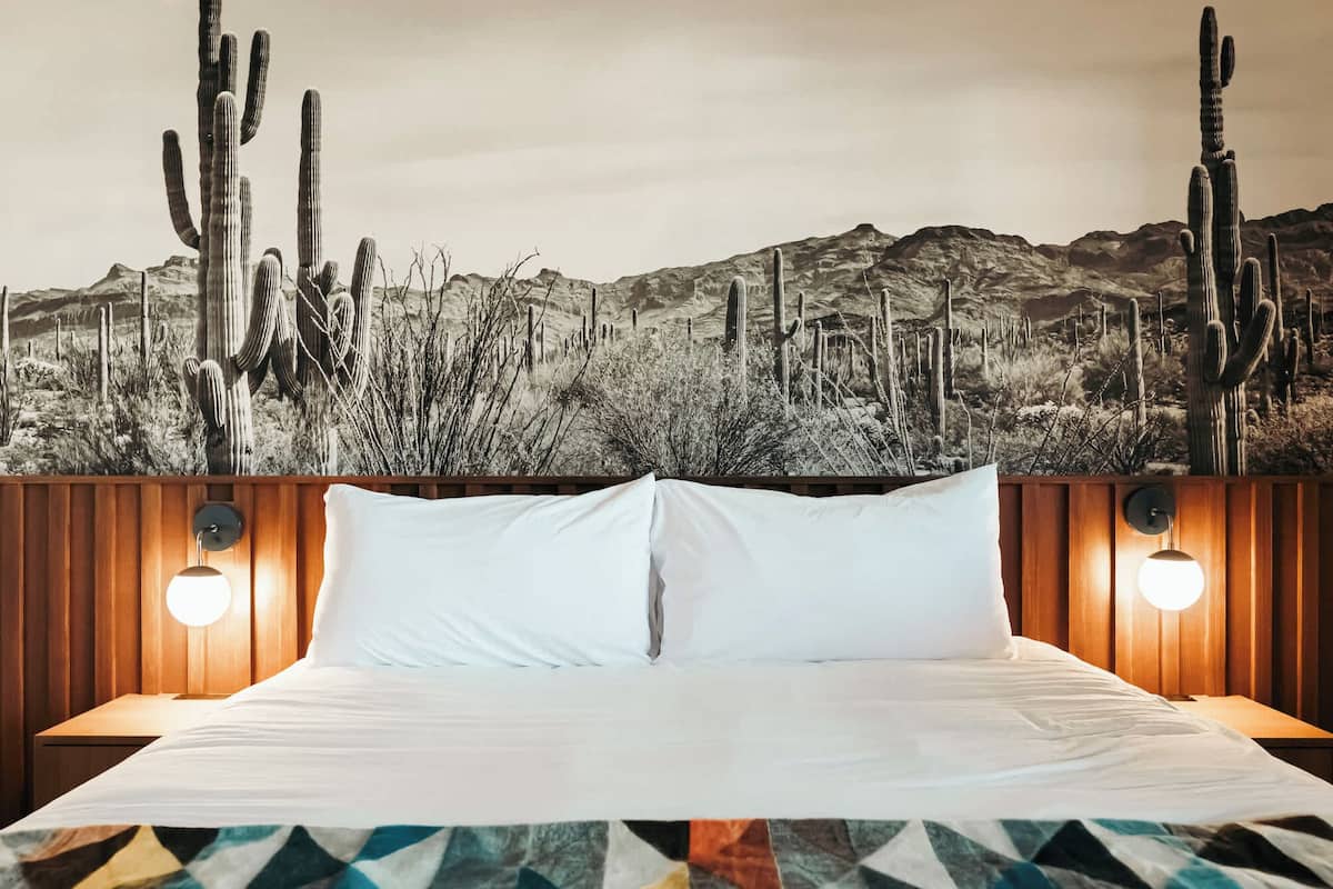 Una cama con sábanas blancas, dos lámparas de noche y un mural con motivos del desierto en la pared.