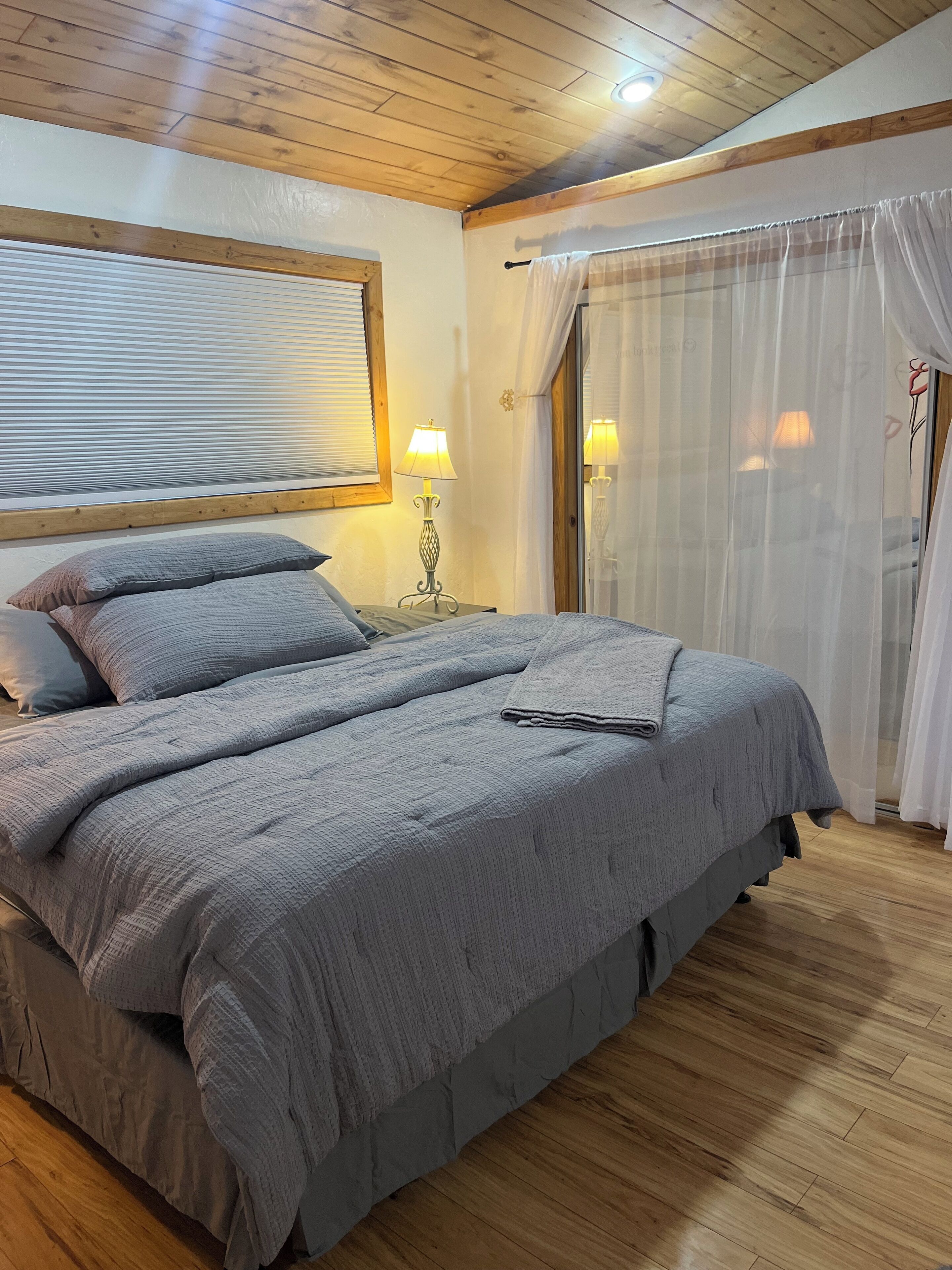 A bedroom with a large bed, wooden ceiling, a nightstand with a lamp, and a window with blinds.