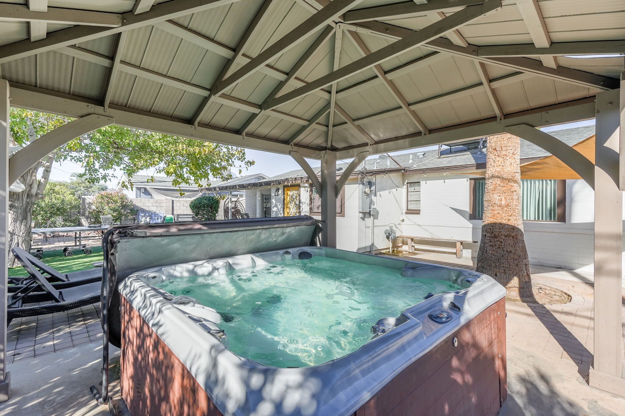 Un jacuzzi bajo un porche cubierto con techo de metal, rodeado por un muro de ladrillo y un árbol.