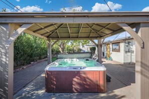Un jacuzzi bajo una pérgola de madera en un patio trasero con un sendero de ladrillos y vegetación.