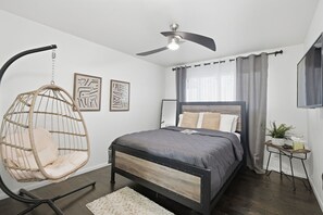 Un dormitorio con cama, una silla colgante, ventilador de techo y una pequeña mesa con una planta.