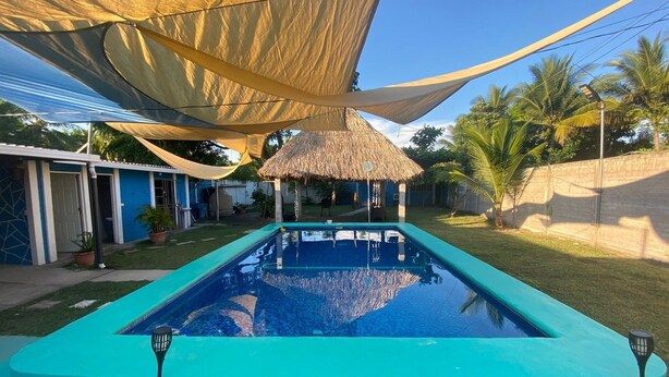 A swimming pool with a thatched-roof structure in the background, surrounded by a blue fence and shaded by a large canopy. - 3-bedroom house in PLAYA LAS HOJAS , SAN PEDRO ,MASAHUAT DPT LA PAZ. (san pedro masahuat)