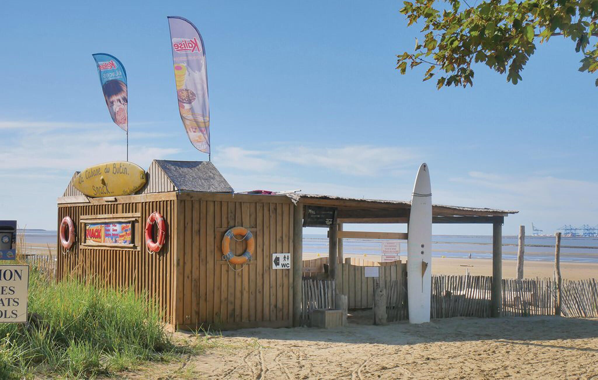 Un petit kiosque de plage où l’on peut acheter des collations, avec une bouée de sauvetage, une planche de surf et un panneau indiquant l’emplacement de la zone de vente de collations.