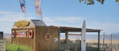 Un petit kiosque de plage où l’on peut acheter des collations, avec une bouée de sauvetage, une planche de surf et un panneau indiquant l’emplacement de la zone de vente de collations.