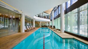 Una piscina interna con acqua cristallina, circondata da pareti di vetro e lettini prendisole in legno.