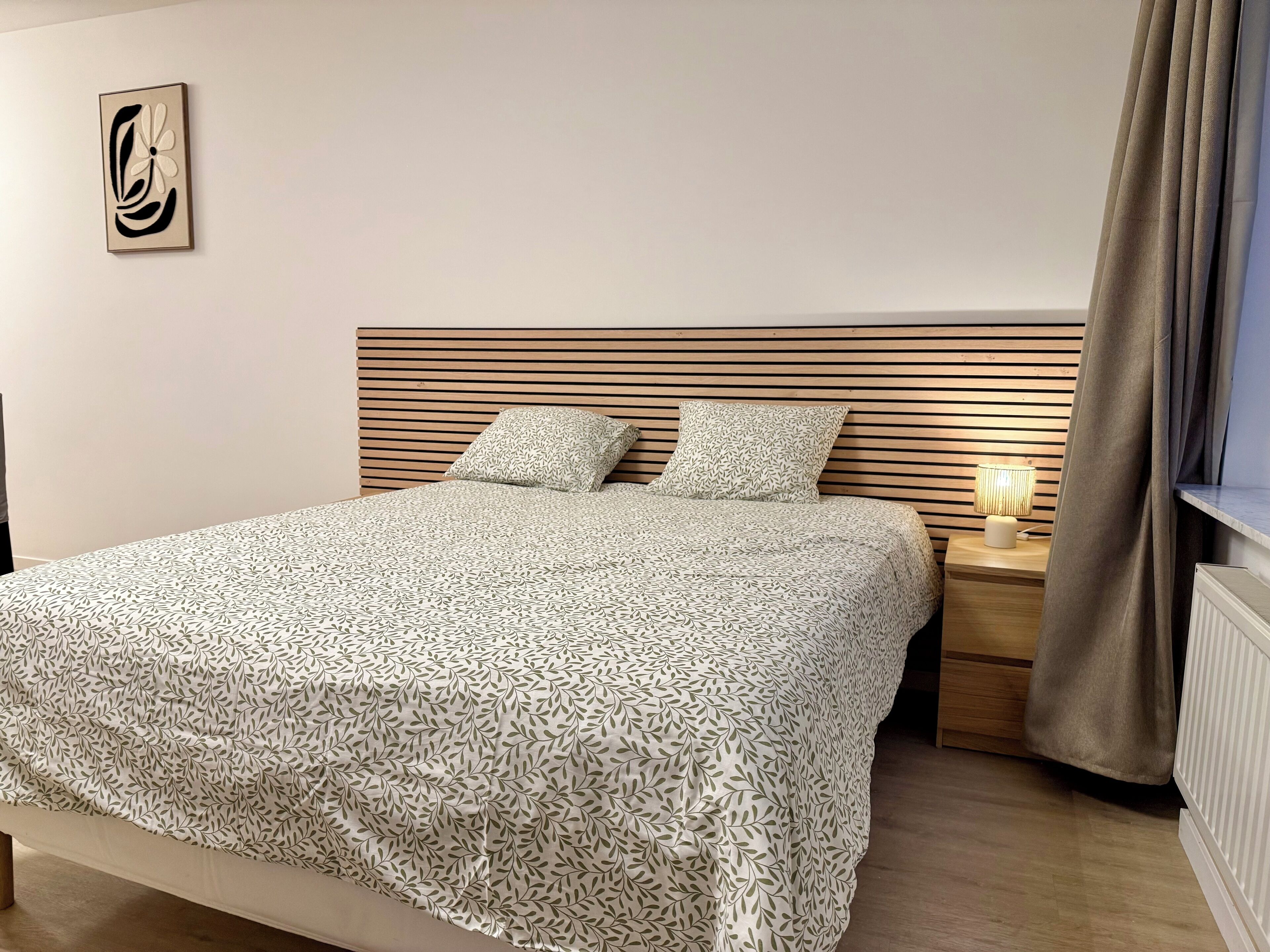 Een net opgemaakt bed met een beddekleed met een patroon, een houten lattenbalken hoofdbord, twee kussens, een bedlampje en een radiator.