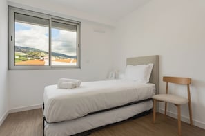 A bedroom with a bed, a chair, a window with a view of a town, and a bedside table with a lamp. - Isabel Place (Câmara de Lobos)