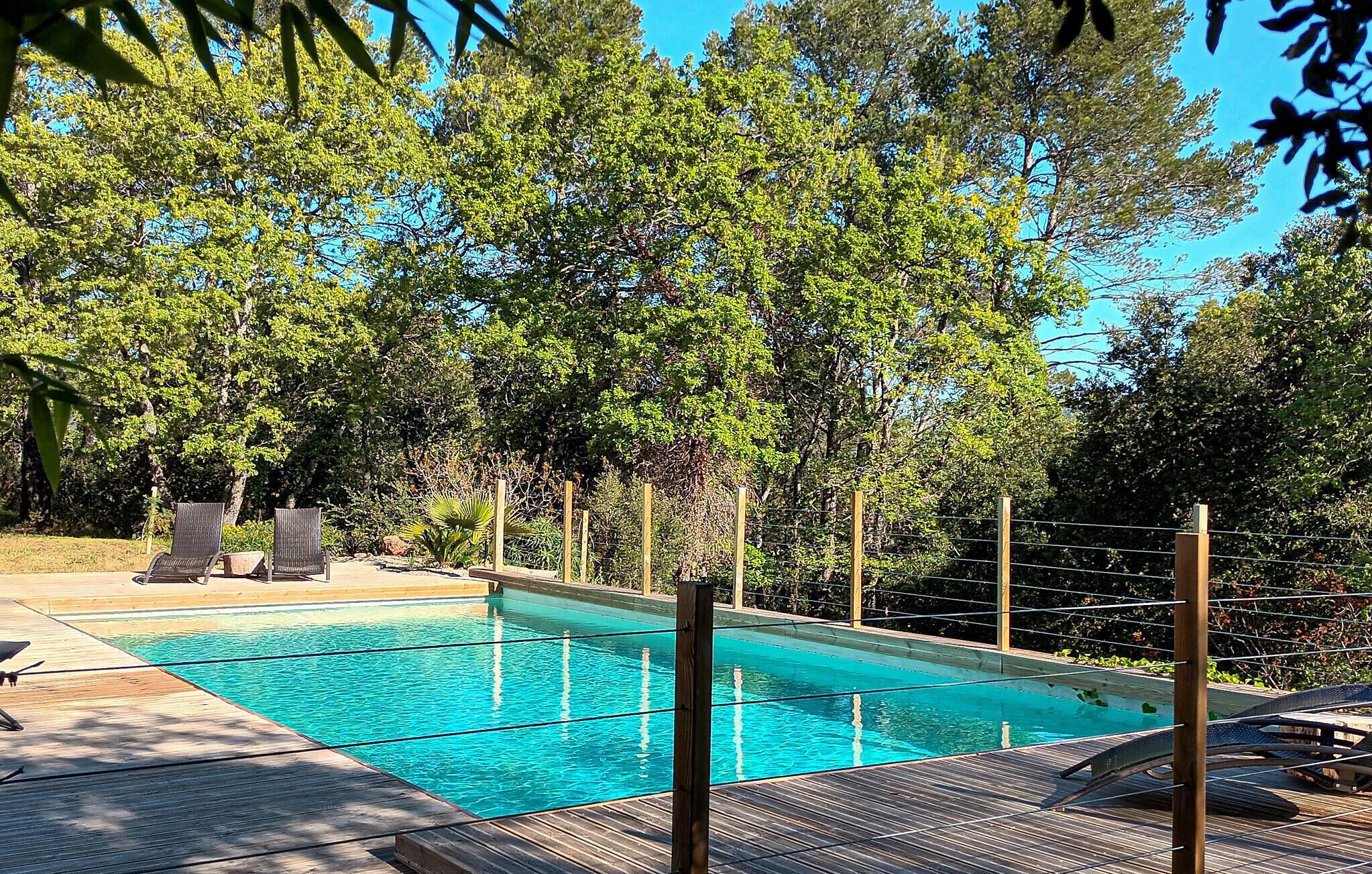 Une piscine entourée d’arbres, avec des chaises longues en bois et une clôture.