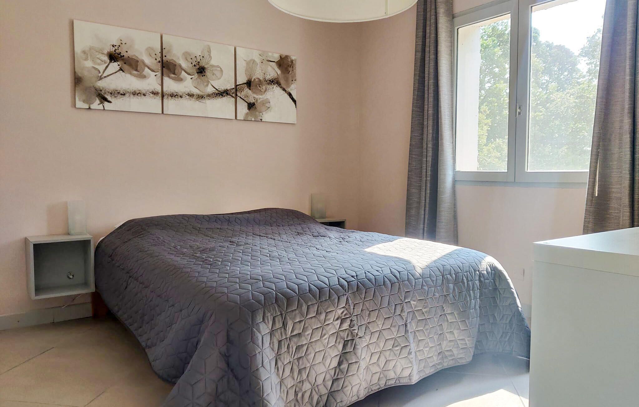 A bedroom with a bed, a nightstand, a bedside table, a window with curtains, and a wall-mounted artwork.