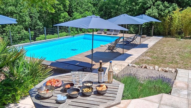 Un espace aménagé au bord de la piscine, avec une table en bois dressée pour un repas, entourée de chaises longues et de parasols.