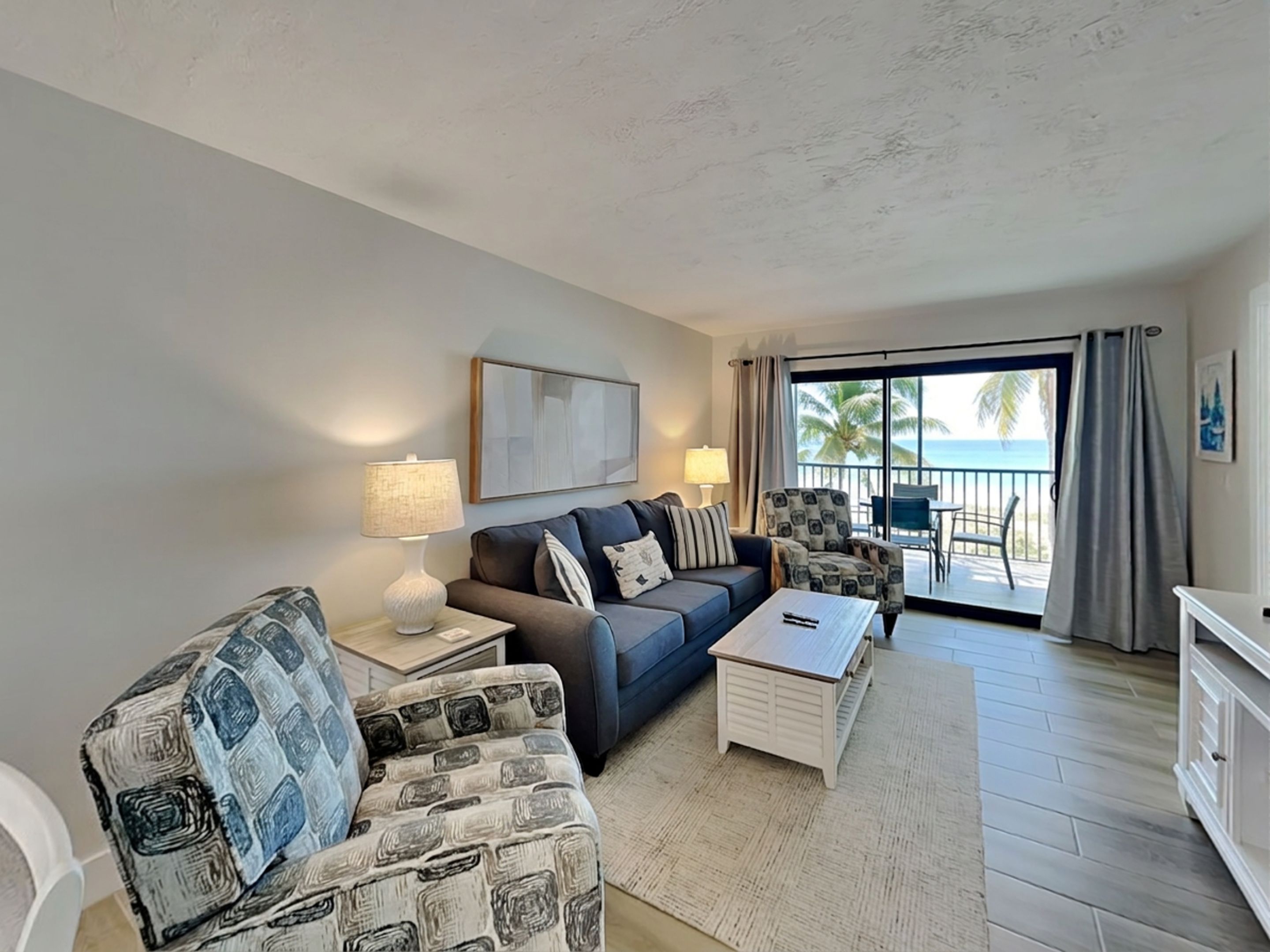 A living room with a sofa, armchair, coffee table, and a view of the ocean through a sliding glass door.