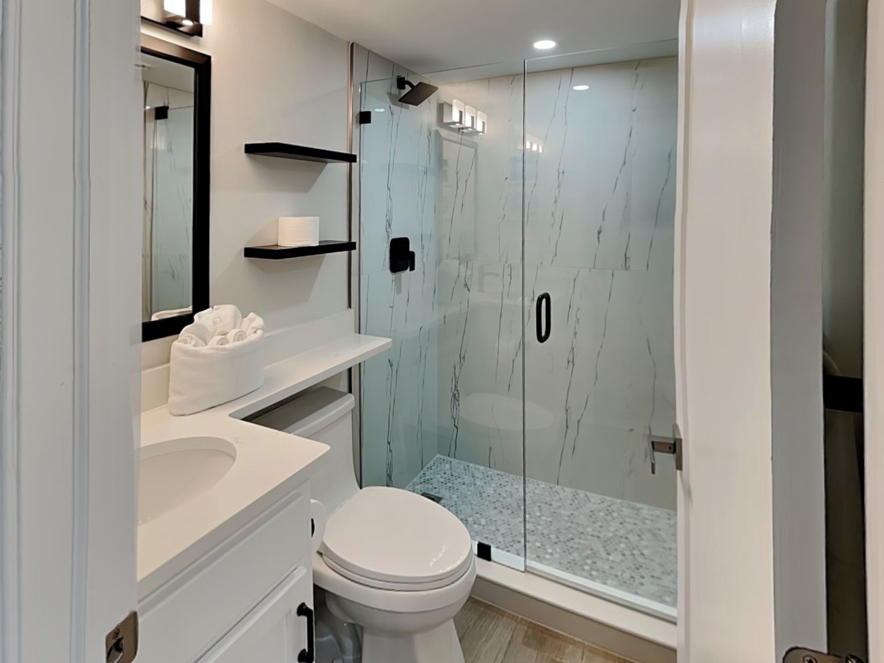 Une salle de bain moderne avec une douche à parois de verre, des toilettes blanches et un meuble-lavabo doté d’un miroir et d’étagères.