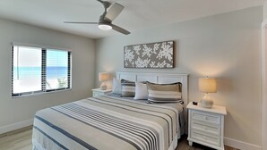 A bedroom with a bed, two bedside tables, a ceiling fan, a window with blinds, and a wall-mounted artwork. - Seaside 209 (Fort Myers Beach)