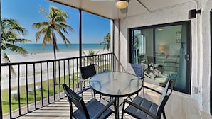 A balcony with a glass table and chairs, a ceiling fan, and a view of the beach and palm trees. - Seaside 209 (Fort Myers Beach)