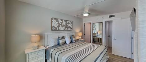 Un dormitorio con cama, mesitas de noche, ventilador de techo y puerta que da a otra habitación.