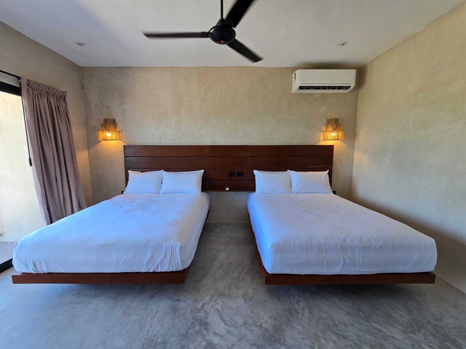 A hotel room with two beds, a ceiling fan, wall-mounted air conditioning, and two wall sconces.