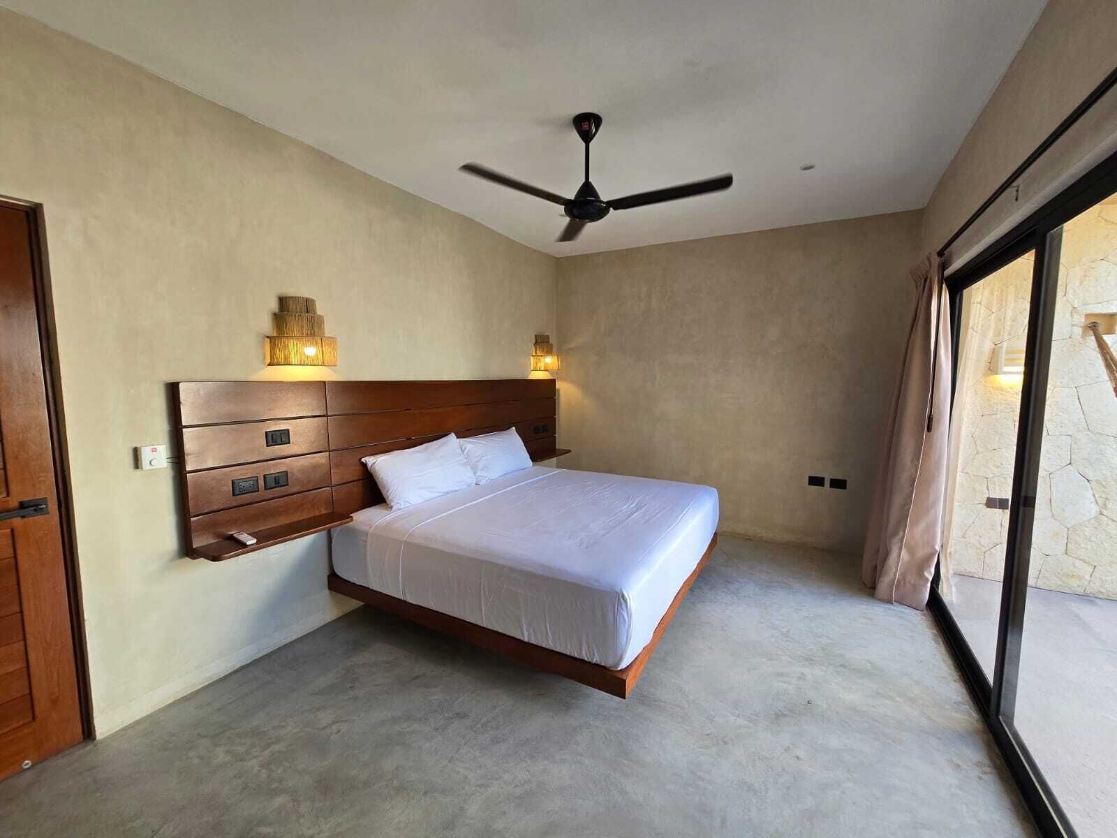 A bedroom with a large bed, a wooden headboard, two wall-mounted lights, a ceiling fan, and a view through a large window.