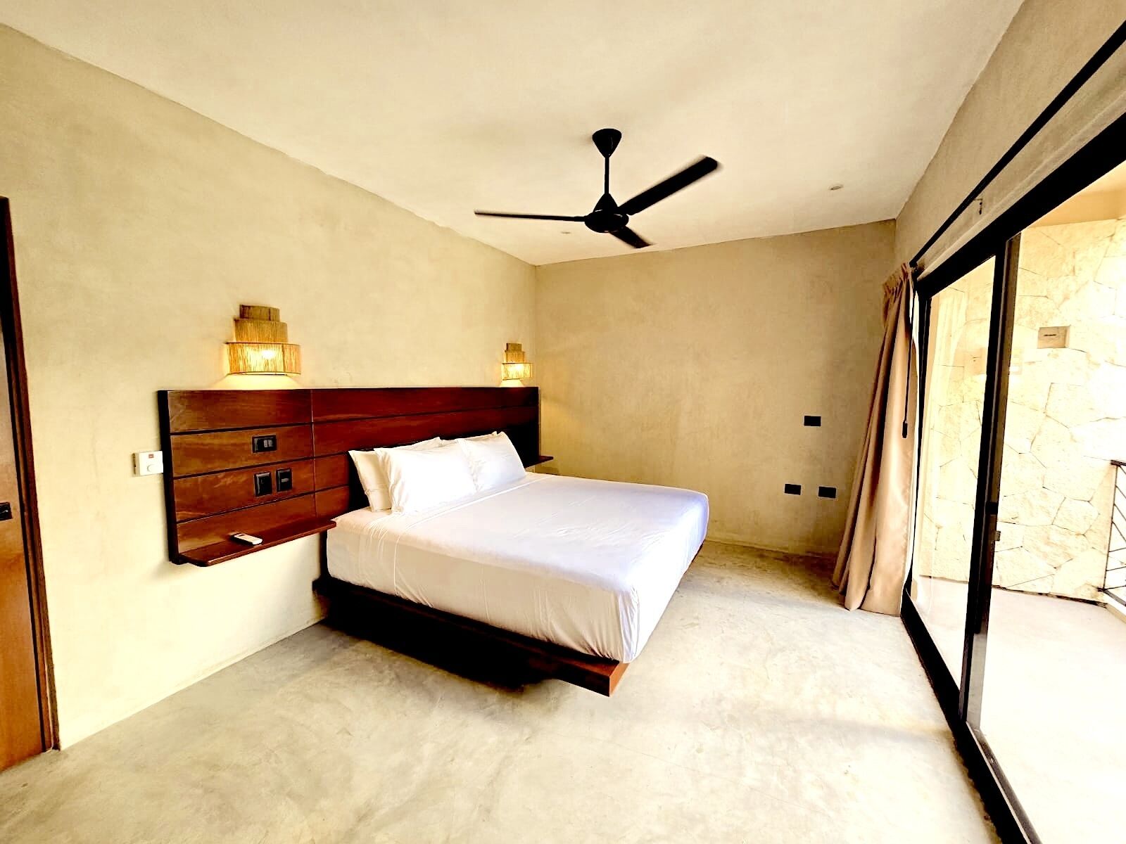 A bedroom with a large bed, a wooden headboard, wall-mounted lights, and a ceiling fan.