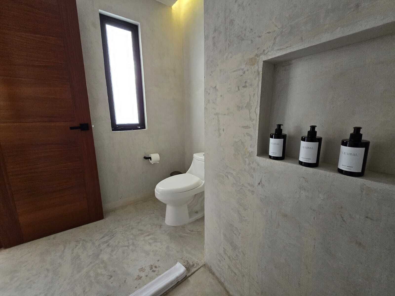 Un baño con inodoro, una puerta de madera y un estante con tres botellas.