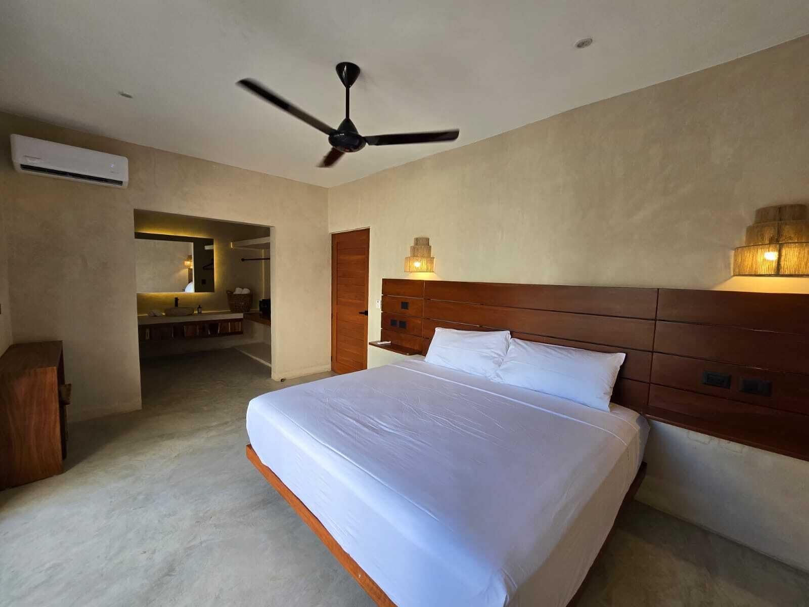 Une chambre d’hôtel avec un grand lit, un ventilateur de plafond, une tête de lit en bois et une salle de bain visible à travers une porte ouverte.