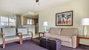 A hotel room with a sofa, armchair, and ottoman, a painting of palm trees, a lamp, and a view of the outside. - HGVC Point At Poipu 2 Bed 2 Bath Unit (Koloa)