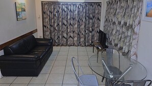 A living room with a black leather sofa, a glass coffee table, a ceiling fan, and patterned curtains. - Cairns holiday unit apartment kitchen aircon wifi near esplanade and hospital (Cairns North)