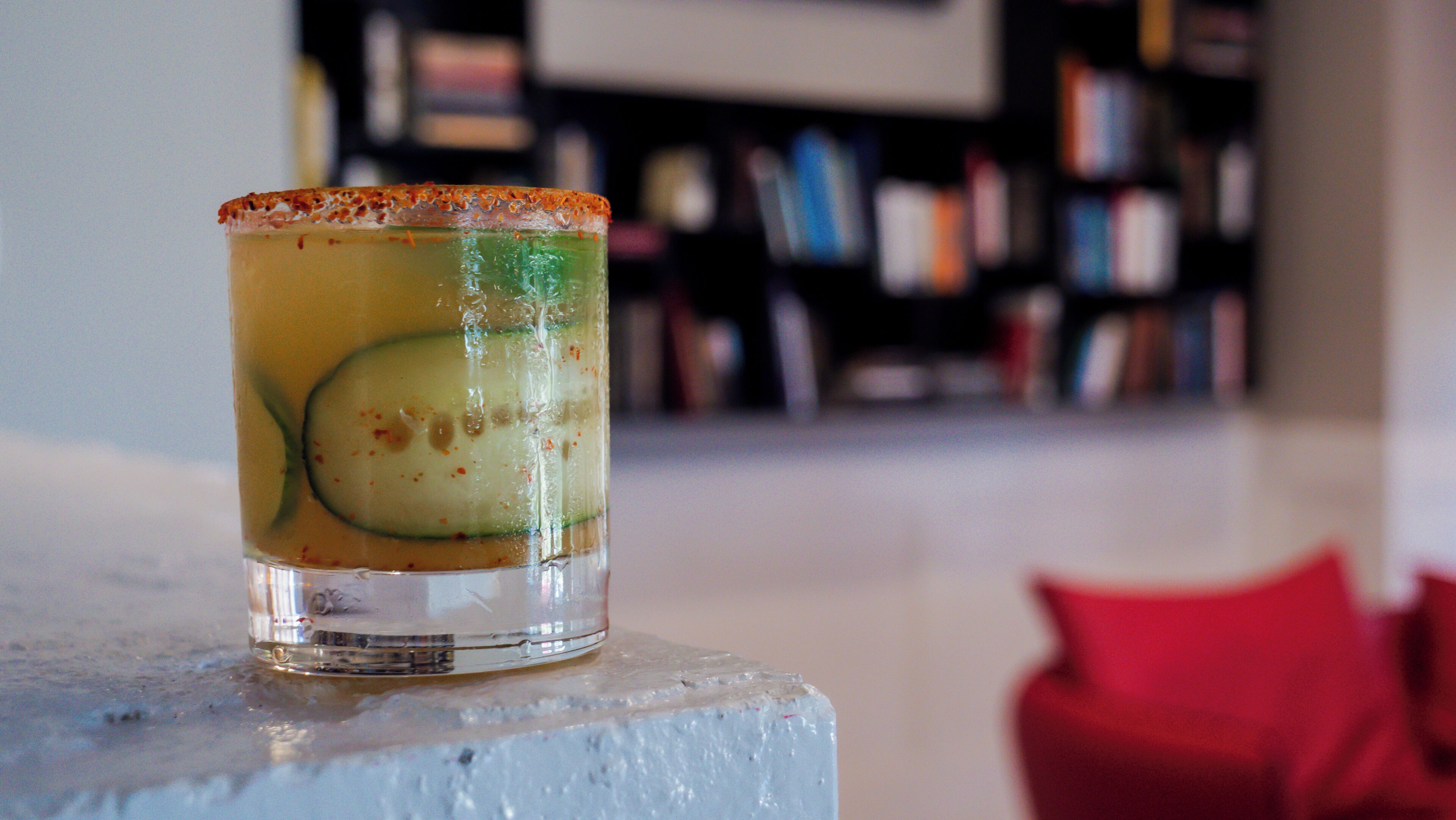 A clear glass with a slice of cucumber and a rim of what appears to be a citrus garnish, placed on a white surface with a blurred background of bookshelves.