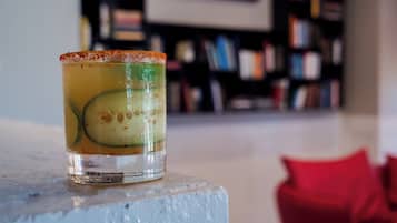 A clear glass with a slice of cucumber and a rim of what appears to be a citrus garnish, placed on a white surface with a blurred background of bookshelves.