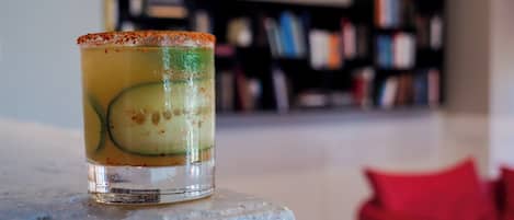 A clear glass with a slice of cucumber and a rim of what appears to be a citrus garnish, placed on a white surface with a blurred background of bookshelves.