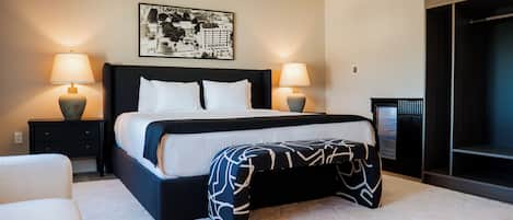 Una habitación de hotel moderna con una cama grande, dos lámparas de noche, una obra de arte con una vista urbana, un armario y un pequeño rincón de descanso.