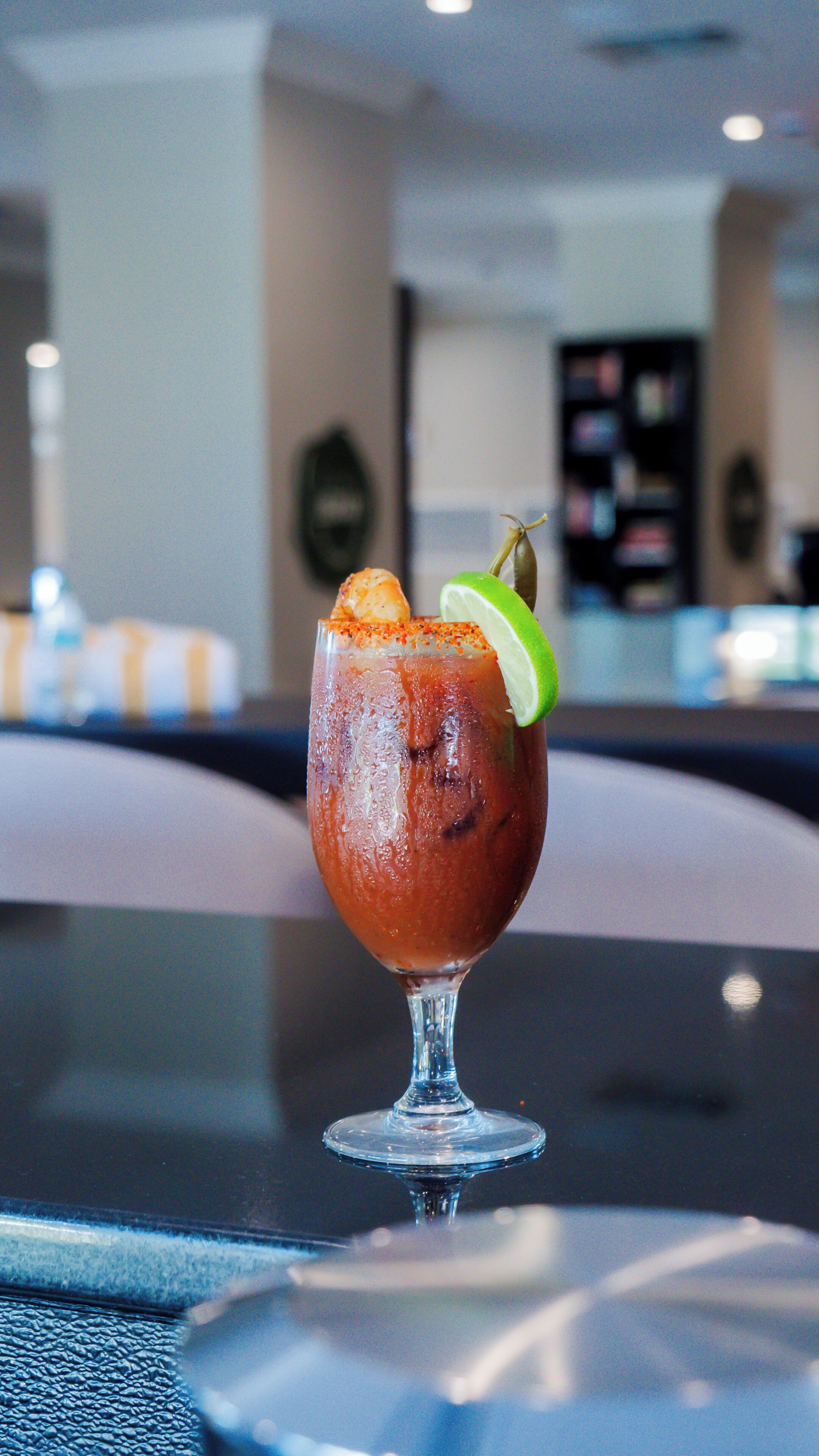 A glass of a red beverage with a lime garnish on a table in a room with a blurred background.
