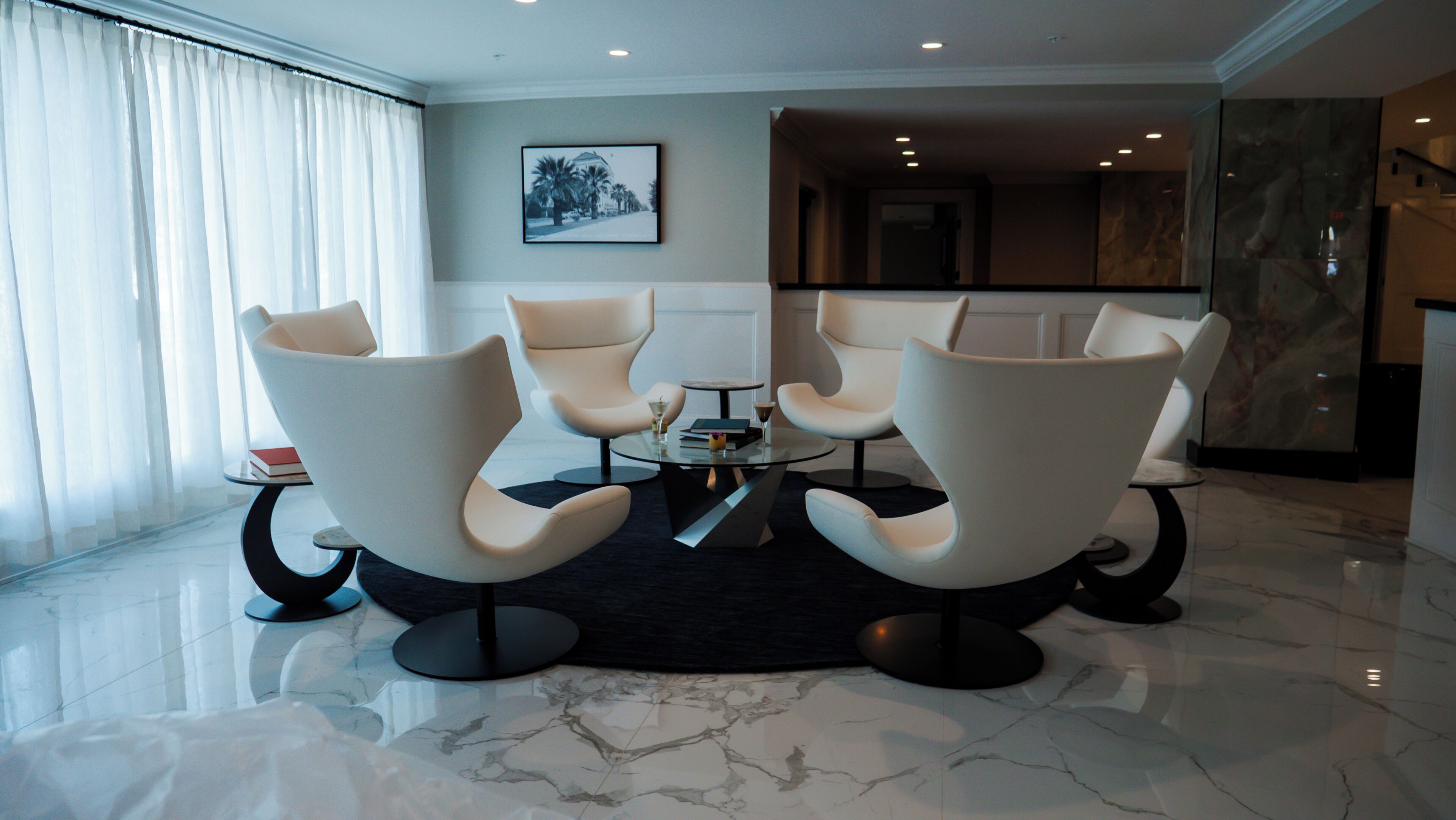 A modern hotel lobby with a marble floor, a round glass table, and four white armchairs arranged around it.