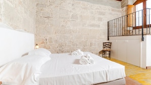 Una cama con sábanas blancas, una silla de madera y una pared de piedra.
