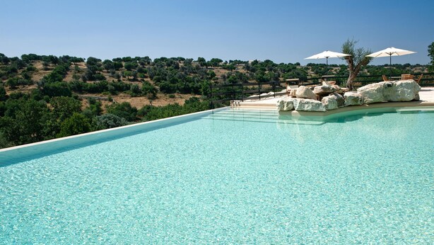 Una piscina infinita azul cristalina con vistas a un paisaje montañoso y mobiliario exterior con sombrillas.