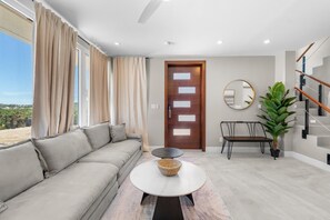 Een moderne woonkamer met een bank, een ronde salontafel, een eettafel met stoelen, een trap en een grote kamerplant.