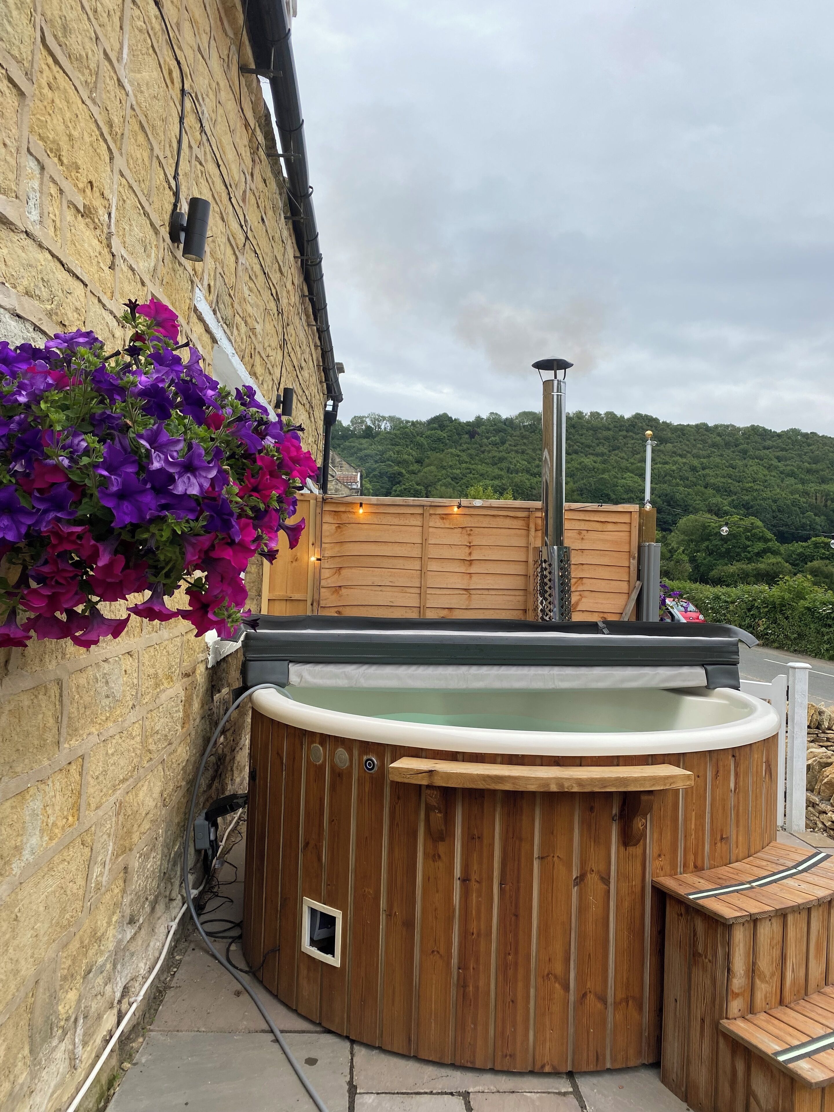 A wooden hot tub with a view of a forested hill, a stone wall, and a flower box with purple flowers.