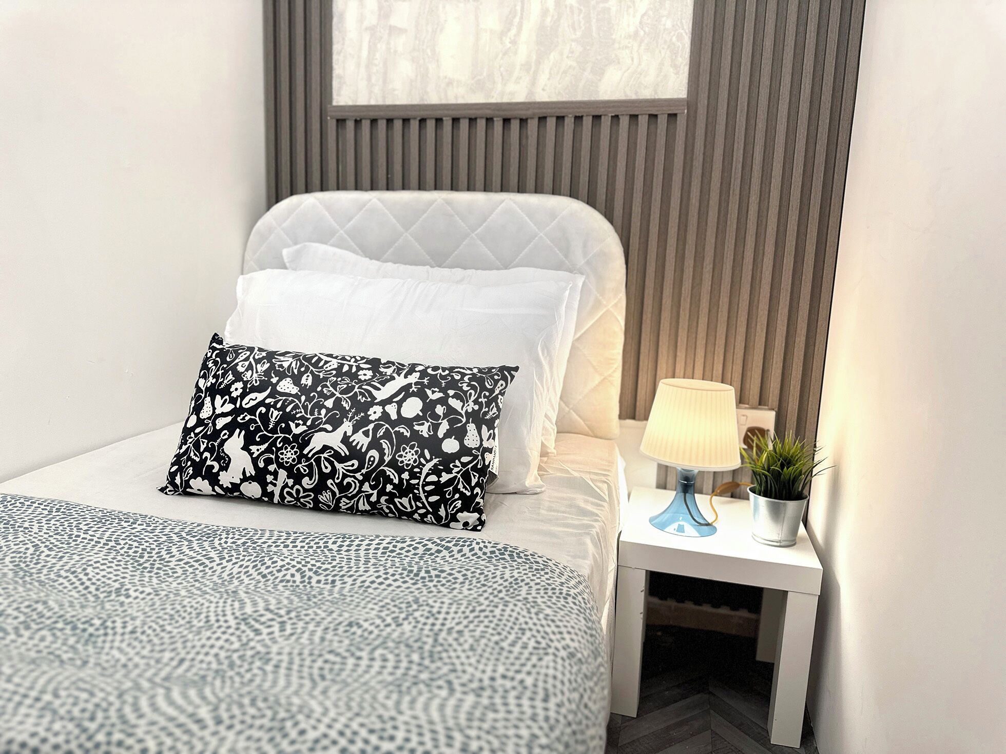 Ein ordentlich bezogenes Bett mit einem gemusterten Überwurf, einem gesteppten Kopfkissen und einem Zierkissen mit einem schwarz-weißen Blumenmuster. Ein Nachttisch mit einer Lampe und einer kleinen Pflanze steht daneben.