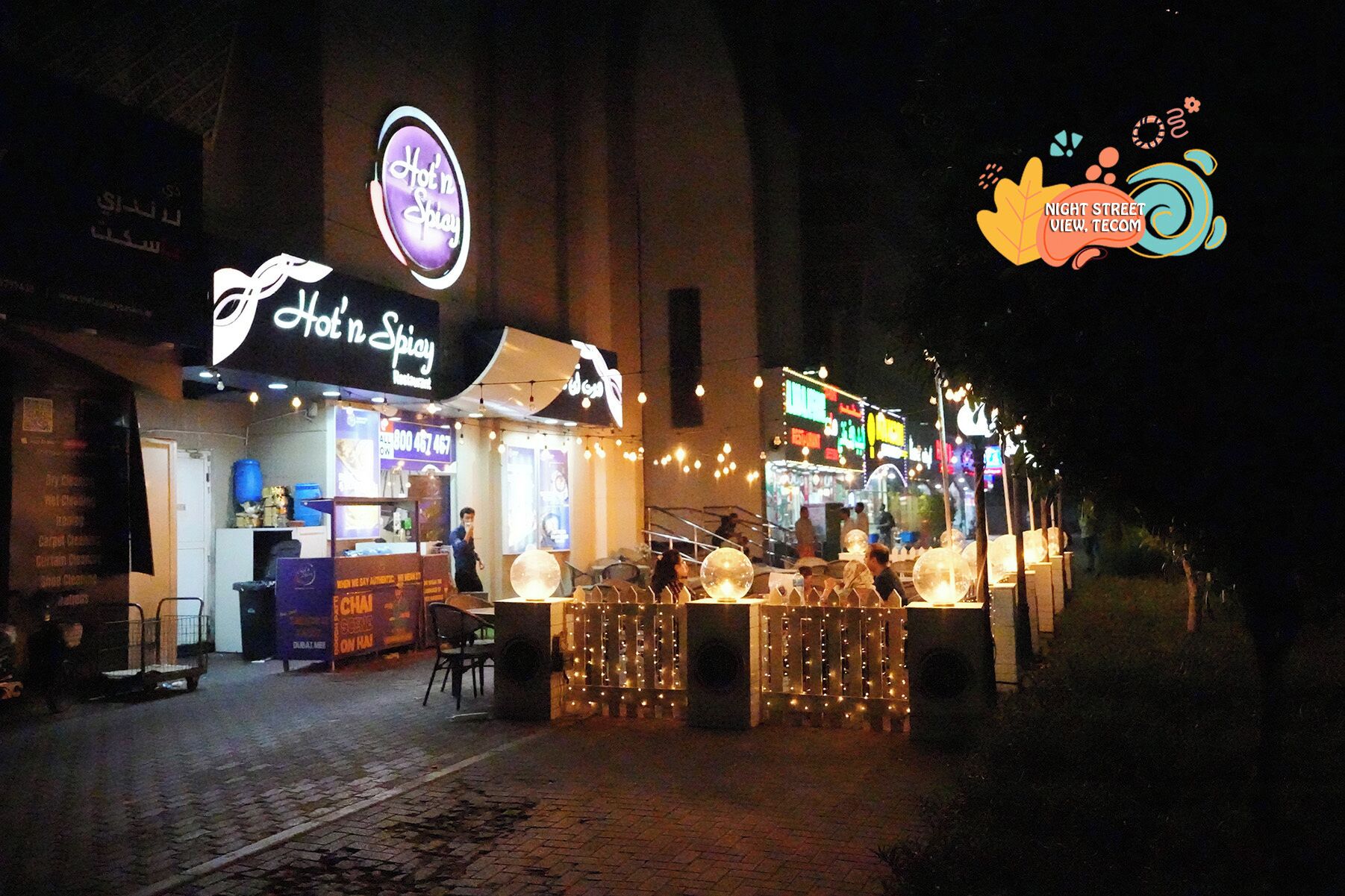 Un puesto de comida callejera con un letrero de neón, mesas al aire libre y luces colgantes.