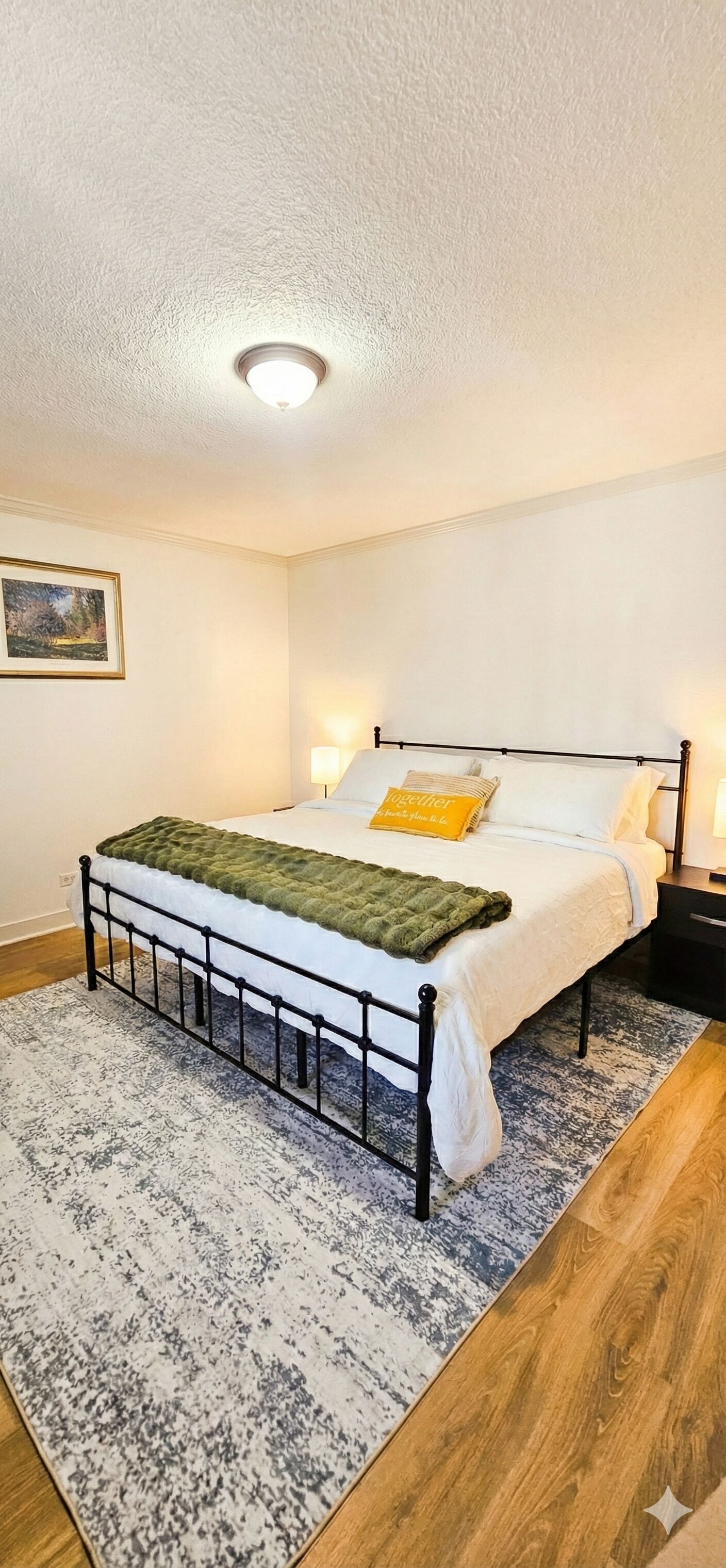 A bedroom with a bed, a nightstand, a lamp, a painting on the wall, and a ceiling light.