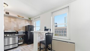 Une cuisine moderne équipée d’appareils électroménagers en acier inoxydable, avec une chaise noire et une vue sur les immeubles par une grande fenêtre.