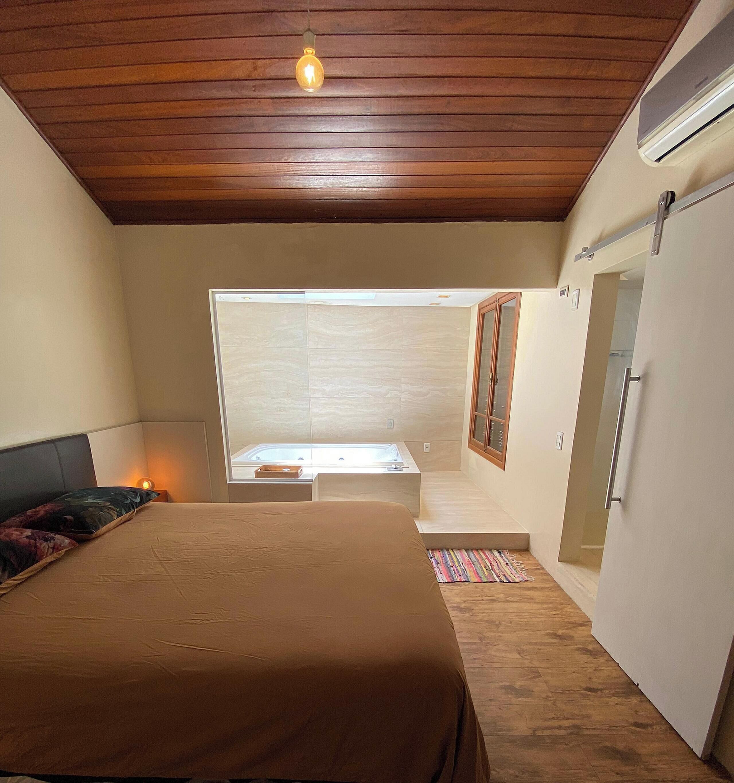 A bedroom with a bed, a small table, a window, and a door leading to a bathroom.