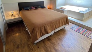 A modern bedroom with a wooden ceiling, a large window, a built-in bench, and a bedside table with a lamp. - Casa 5 Ruas com Hidro - Rio Tavares Florianopolis (Florianópolis)