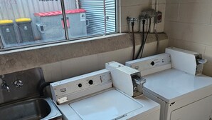 A laundry room with two white washing machines, a stainless steel sink, and a window overlooking a fenced area with recycling bins. - Cairns holiday unit apartment kitchen aircon wifi near esplanade and hospital (Cairns North)