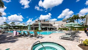 Un área de piscina con sillones de descanso y sombrillas, un edificio grande al fondo y un cielo azul despejado.