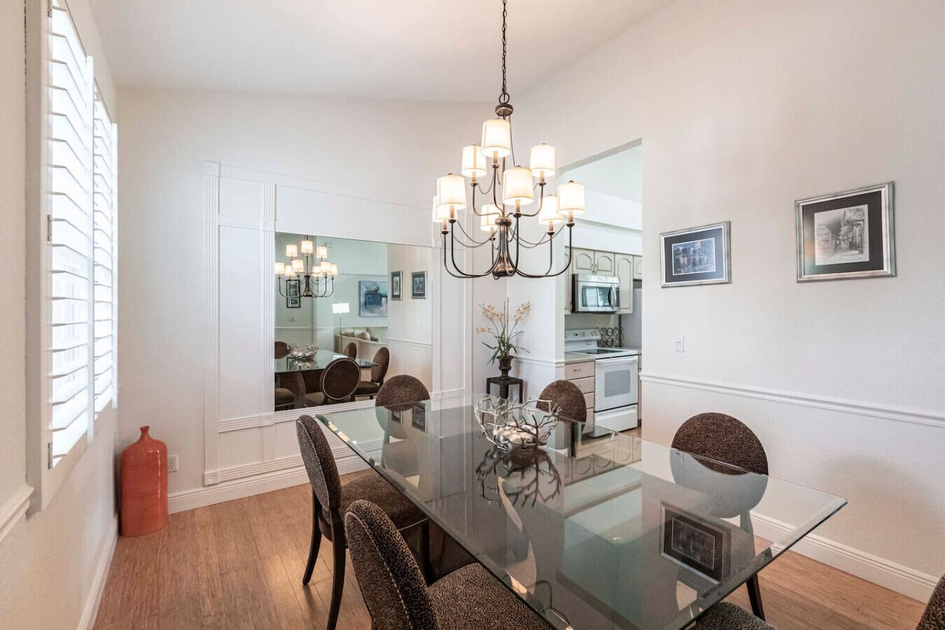 A modern dining room with a glass table, dark chairs, a chandelier, and a kitchen in the background.